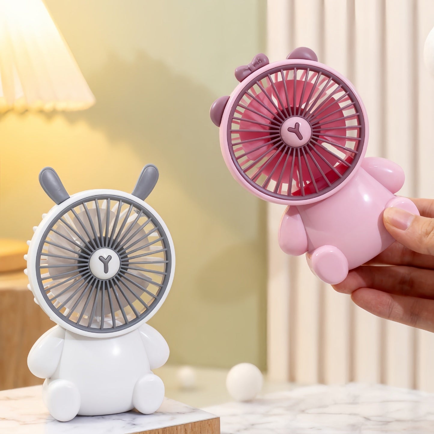 Two cute animal-shaped fans, one pink and one white, on a light-colored surface with a blurred background.