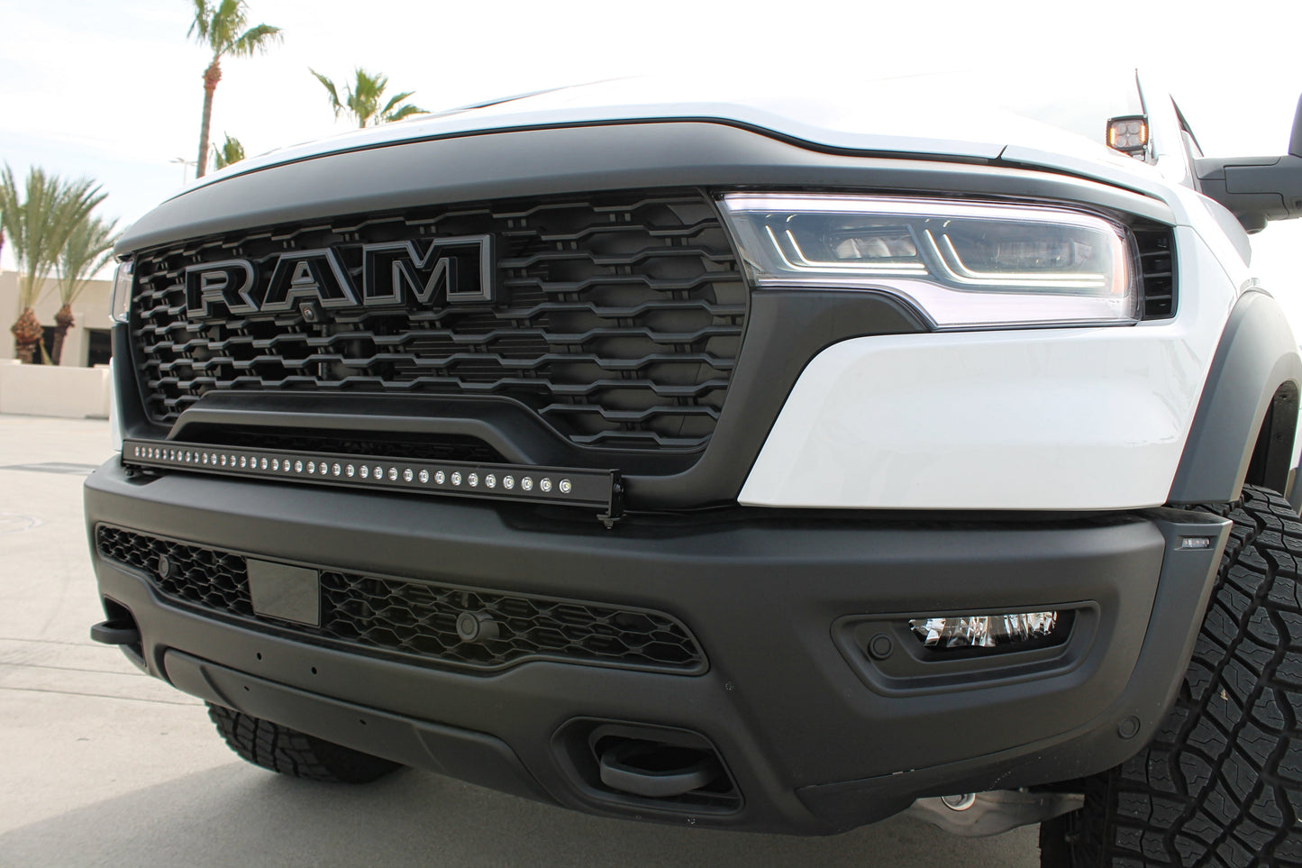 Close-up of a white Ram truck's front grille with light bar and palm trees in the background.