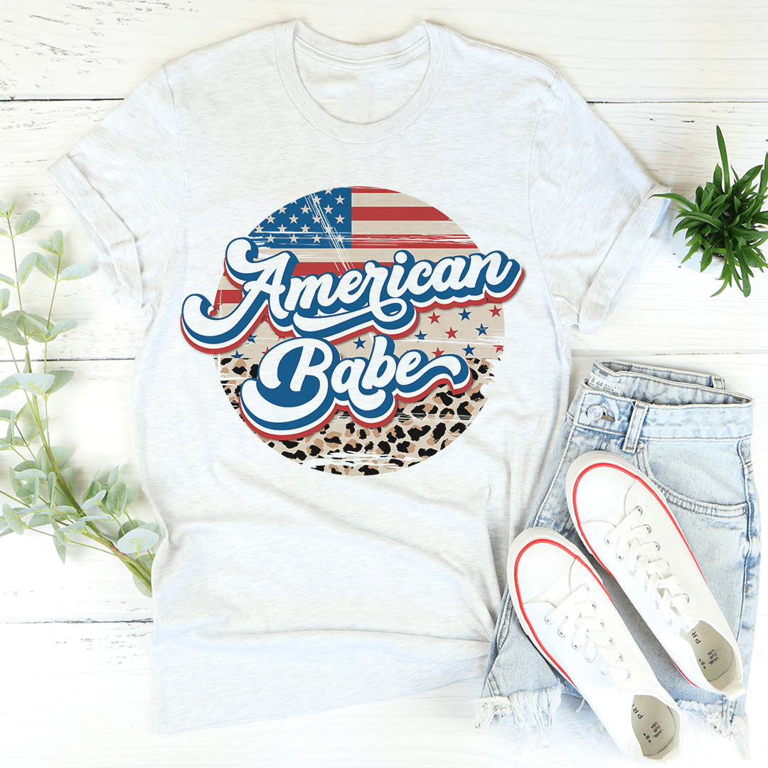 White t-shirt with 'American Babe' design, light blue jeans, and white sneakers on a wooden surface.