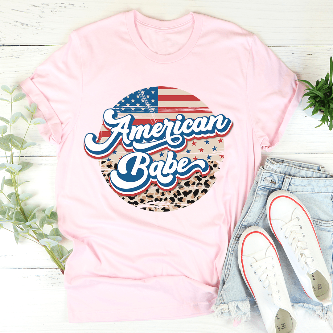 Pink t-shirt with 'American Babe' design on a white wooden surface with jeans and shoes.
