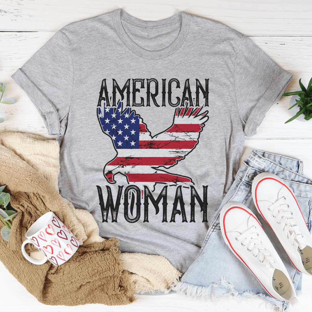 Gray t-shirt with 'American Woman' design featuring an eagle, surrounded by casual items on a light wooden surface.