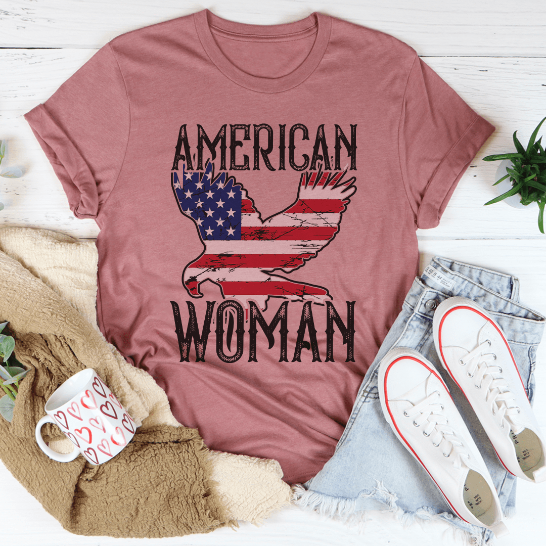 Pink t-shirt with 'American Woman' graphic on a white surface with casual items.