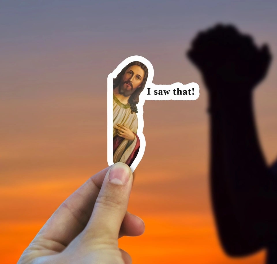Hand holding a sticker with Jesus and 'I saw that!' text against an orange sunset sky.