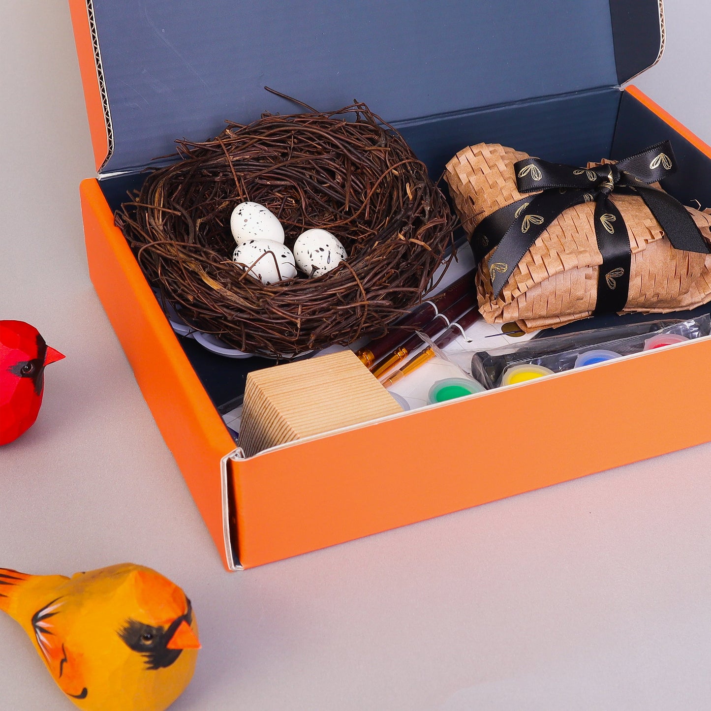Open box containing a nest with eggs, wrapped items, and art supplies on a light gray background.