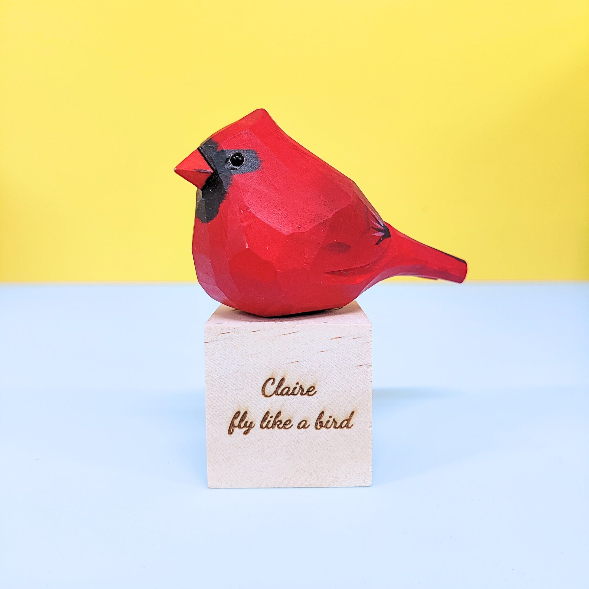 Red cardinal figurine on a wooden block with 'Claire fly like a bird' text against a yellow and blue background