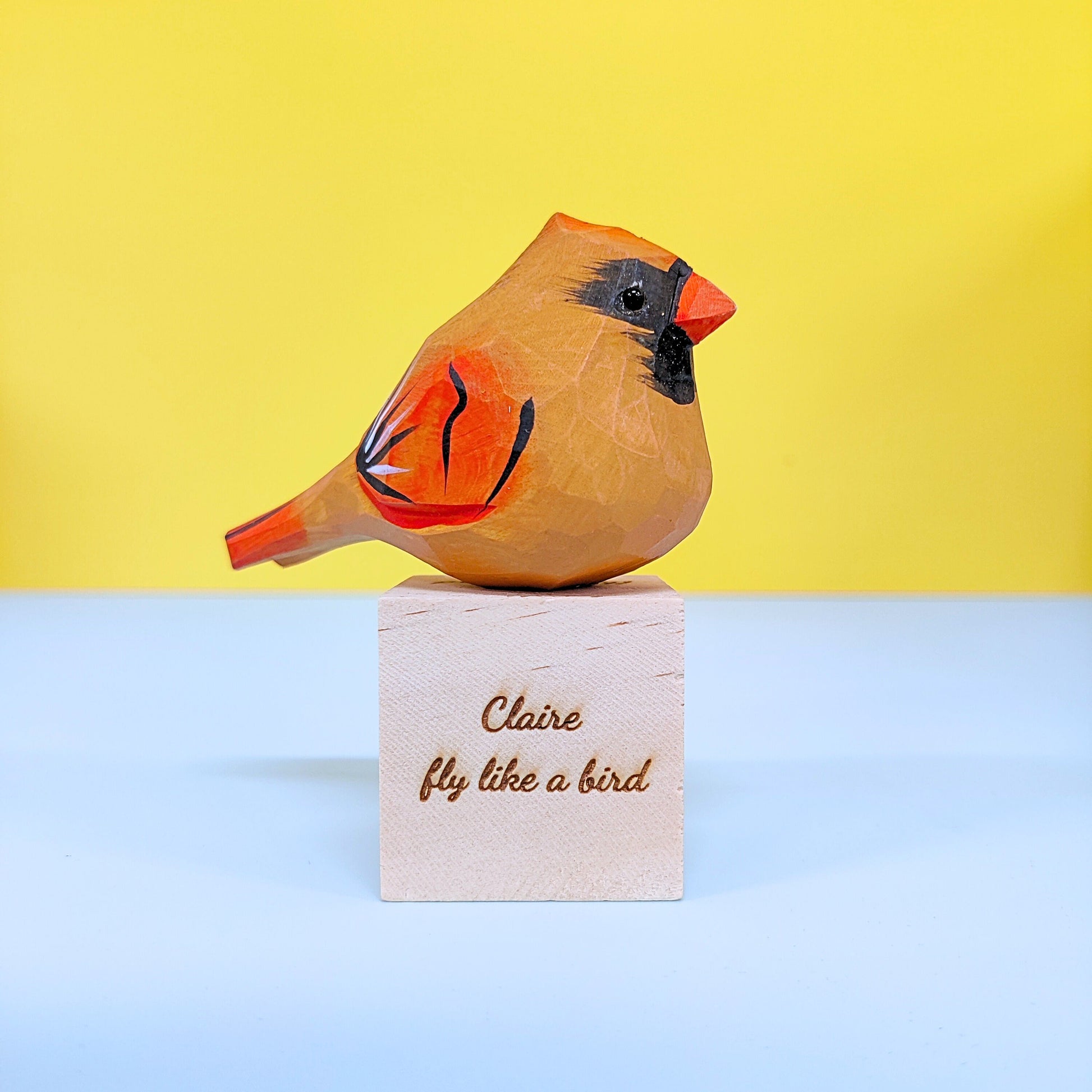 Wooden bird figurine on a block with 'Claire fly like a bird' text against a yellow background