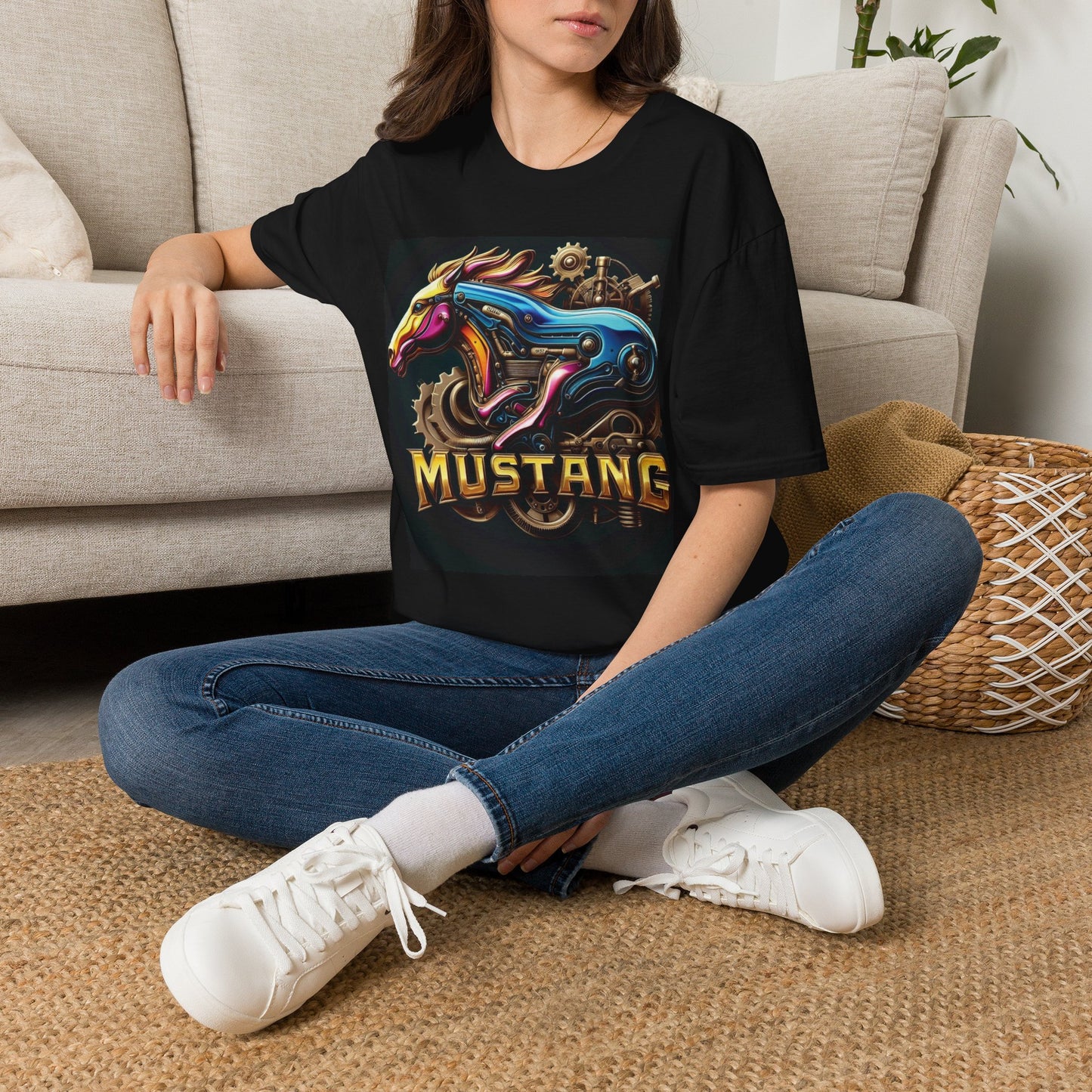 A woman sitting cross-legged on a woven rug in front of a beige sofa, wearing a black graphic T-shirt with a colorful Mustang horse design and the word 'MUSTANG' on the front, paired with blue jeans and white sneakers.