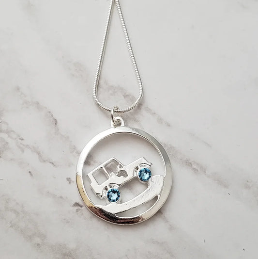 Silver necklace with a circular pendant featuring a Jeep design and blue stones on a marble surface.