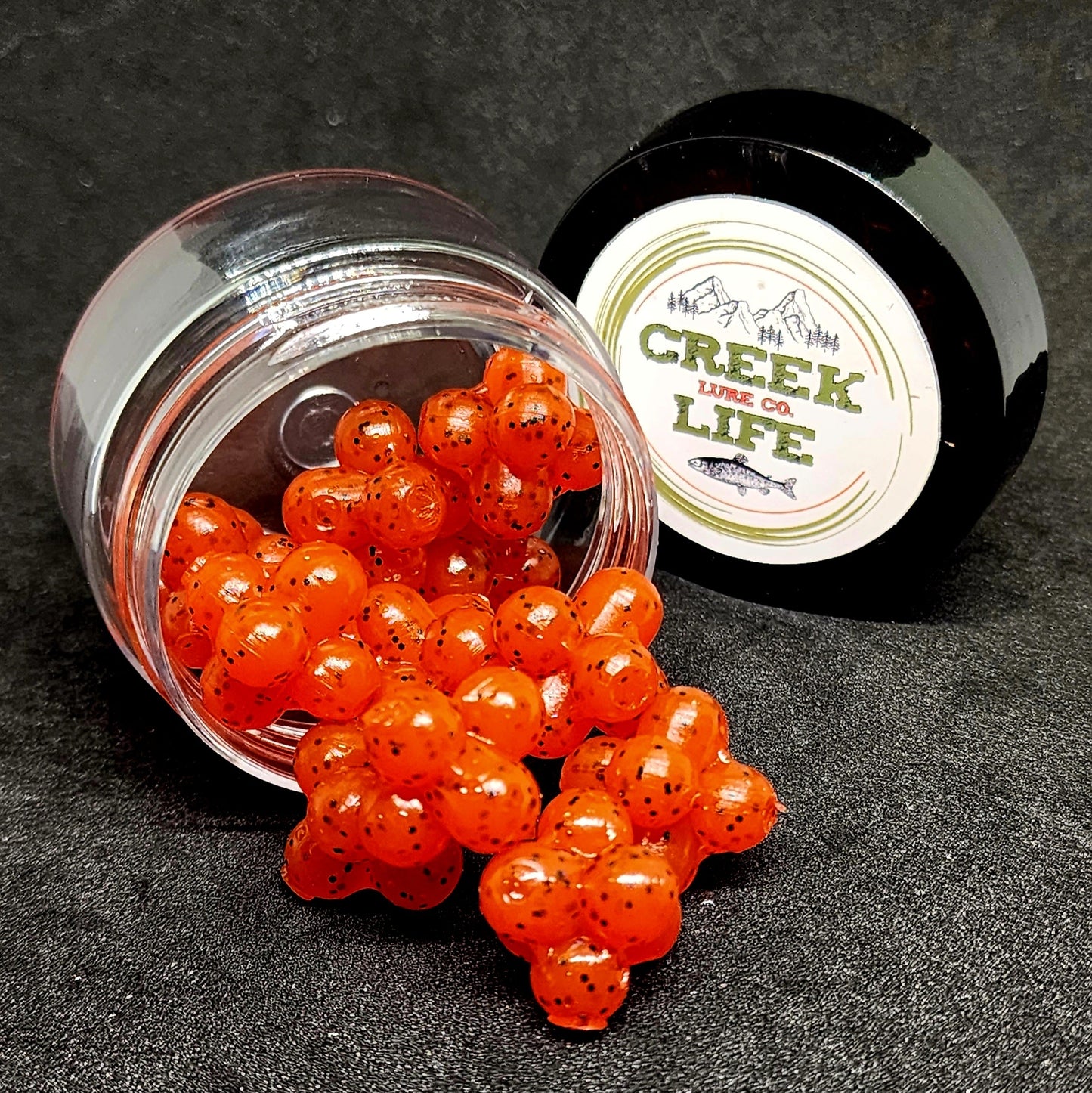Orange fish eggs spilling out of a glass jar with a black lid labeled 'Creek Life Co.' on a dark background.