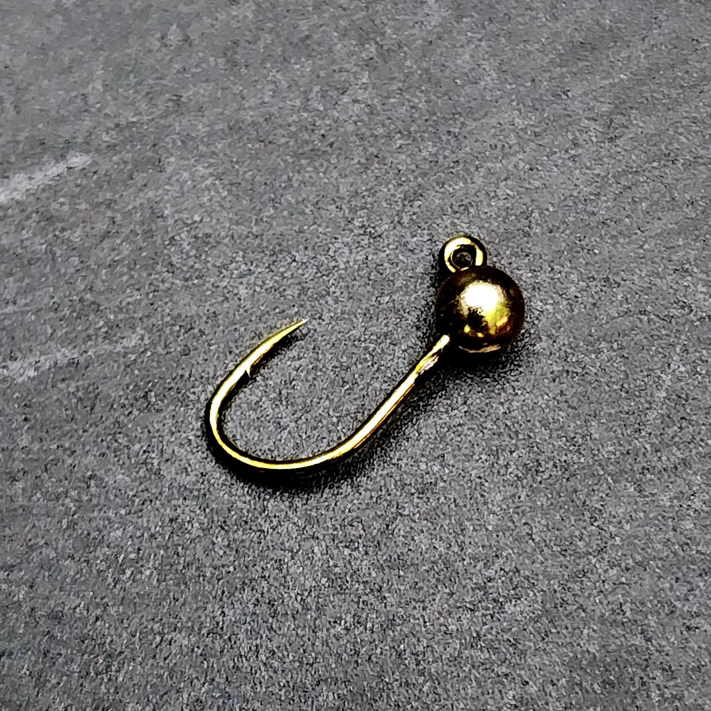 Gold-colored fishing hook jig head on a gray surface