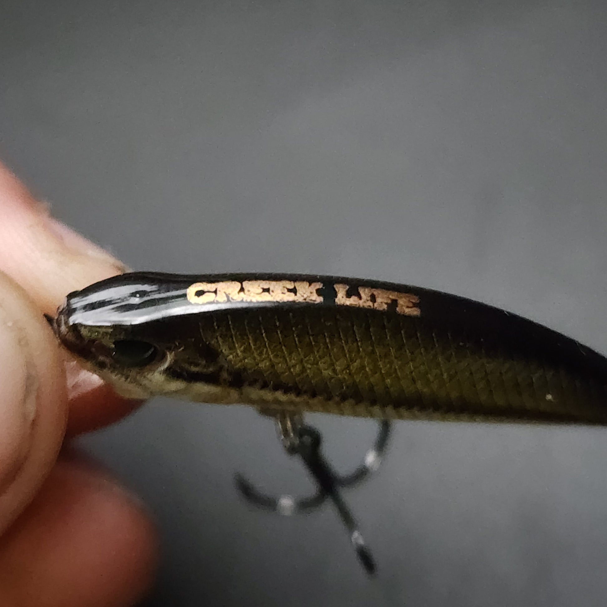 Fishing lure with 'Creek Life' branding held by a hand against a gray background