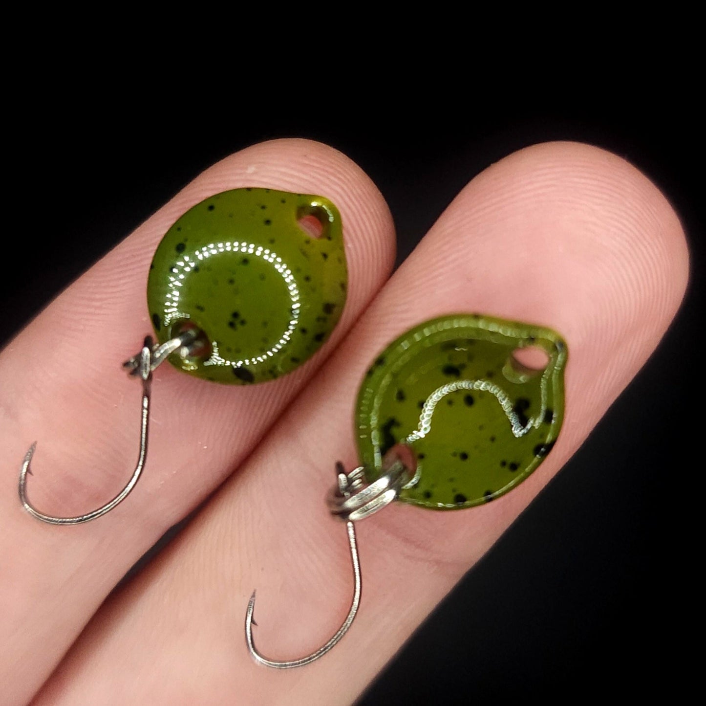 Two green fishing lures with black speckles on a person's hand against a black background.