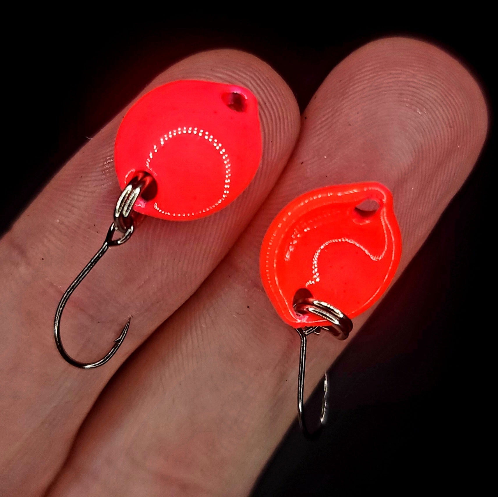 Two red fishing lures with hooks on a person's finger against a dark background