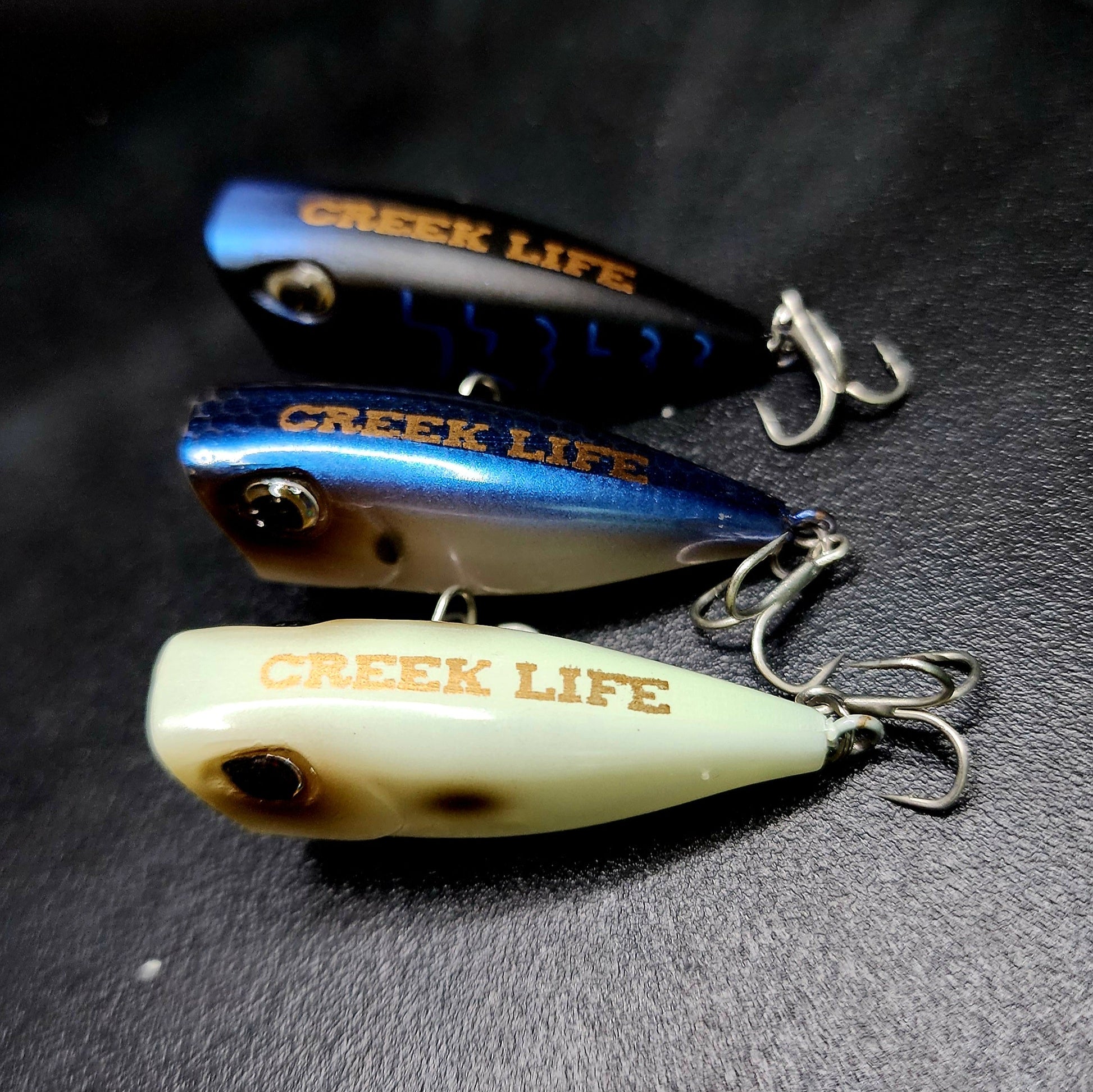 Three 'Creek Life' fishing lures on a dark surface