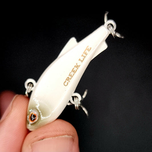 Fishing lure with 'Creek Life' branding held in a hand against a black background