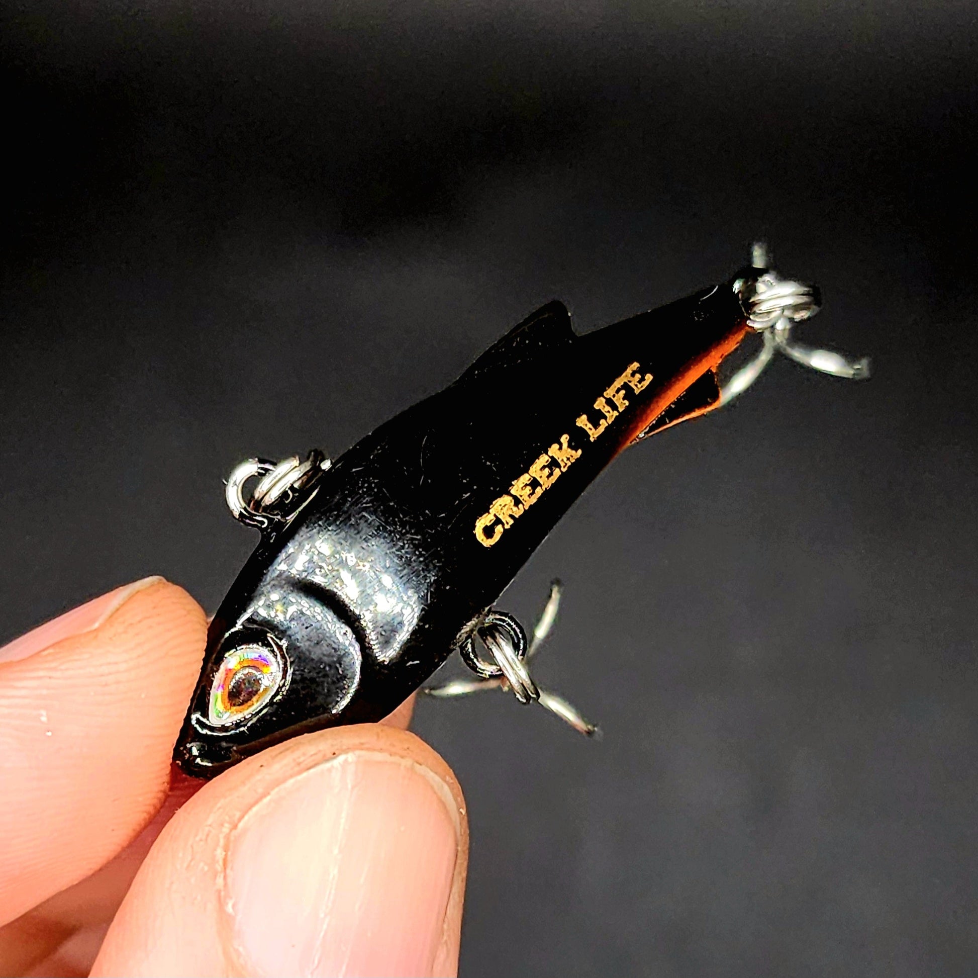 Fishing lure with 'Creek Life' branding held in a hand against a dark background