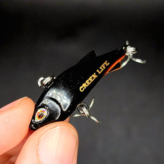 Fishing lure with 'Creek Life' branding held in a hand against a dark background