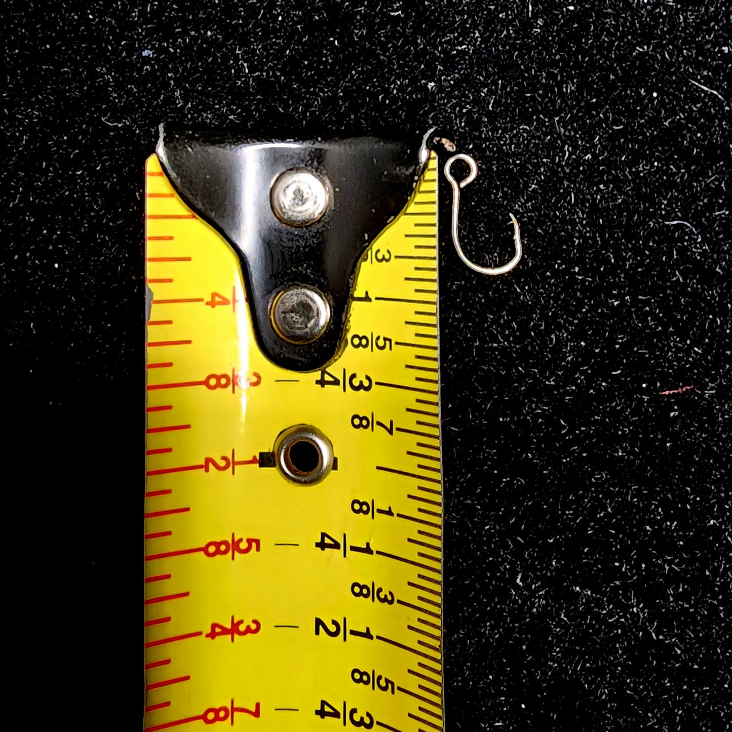 Fishing hook next to a yellow measuring tape on a black background
