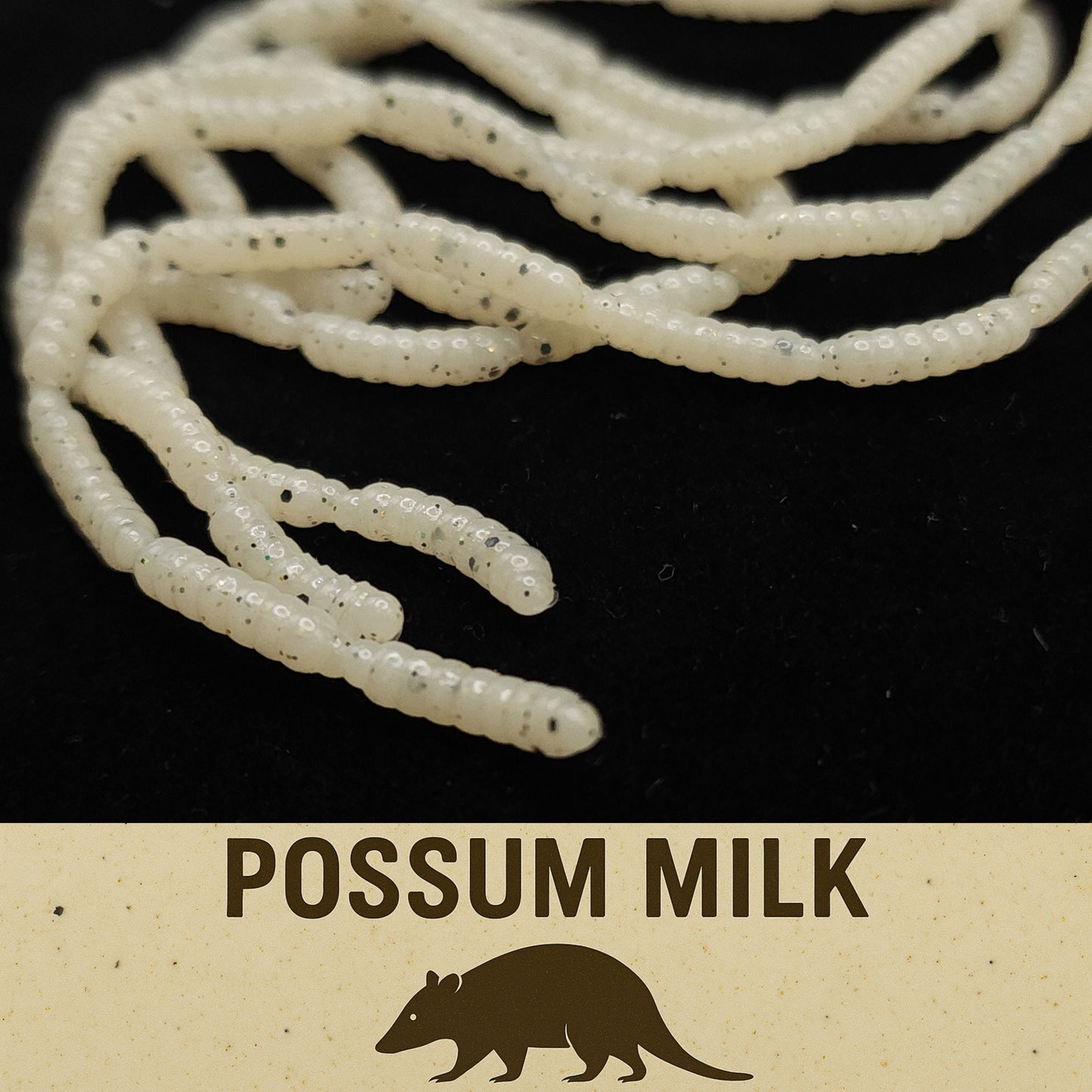 White Micro Maggot Fly Larvae fake bait with black specks on a dark background, labeled 'possum milk'.