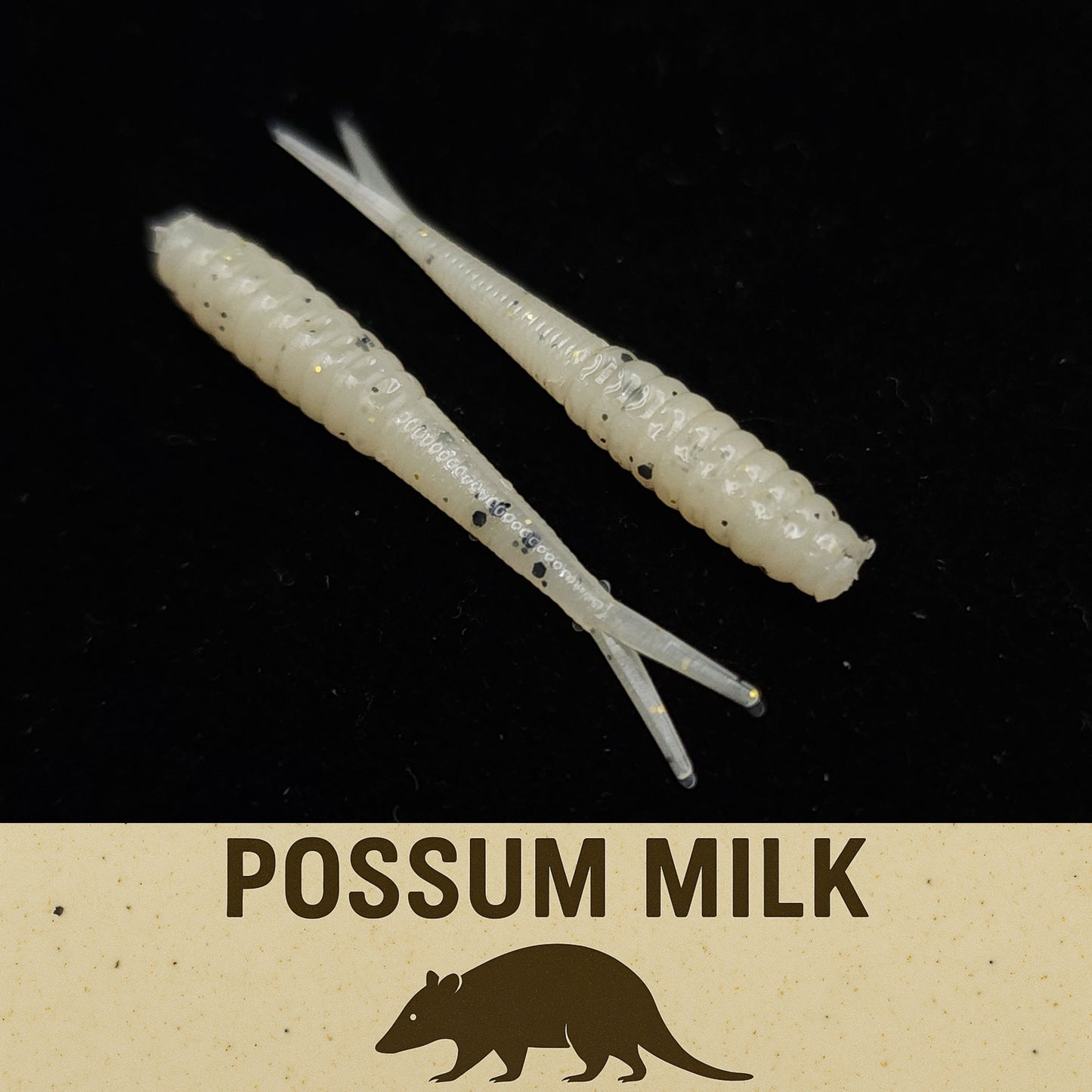 Two white fishing lures with black specks on a black background, labeled 'Possum Milk'.