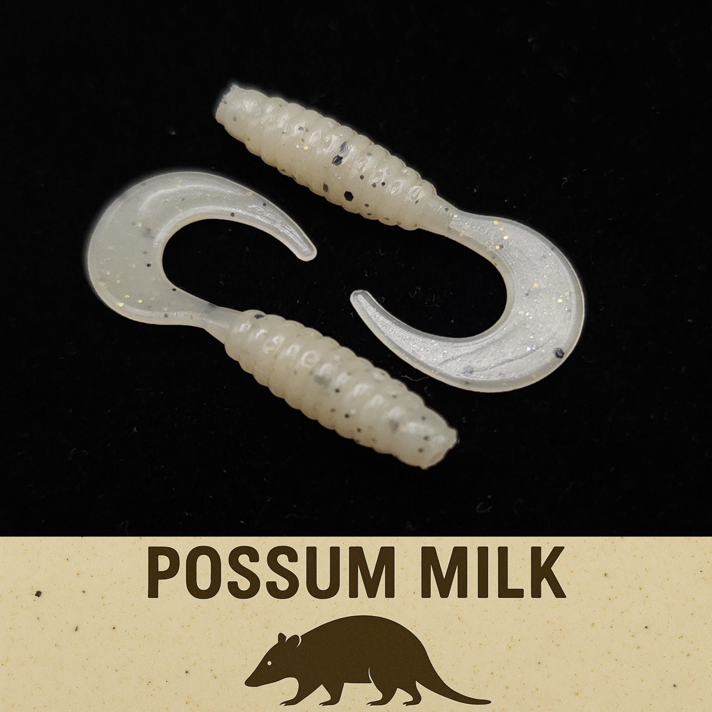 Two white fishing lures with black speckles on a dark background, labeled 'Possum Milk'.