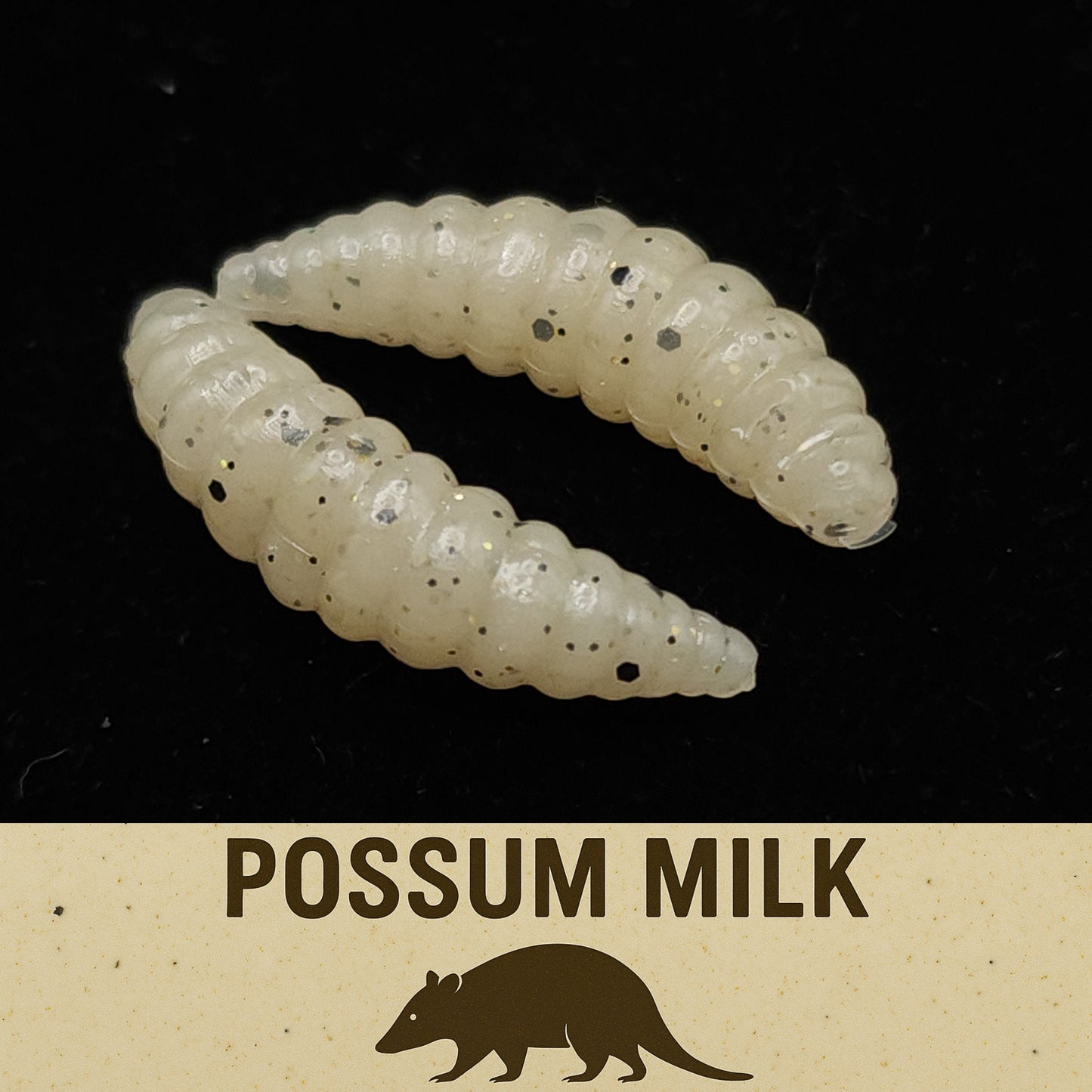 Two white larvae fishing lures with black specks on a black background, labeled 'Possum Milk'.