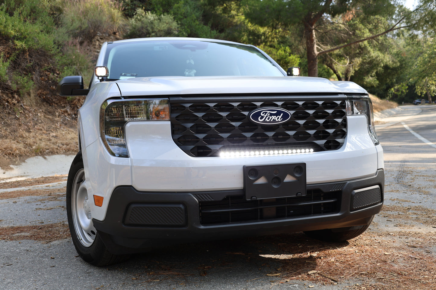 White Ford vehicle with a prominent grille and light bar on a road with trees in the background