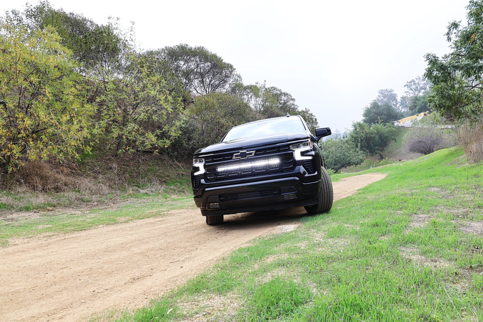 Black Chevrolet truck driving on a dirt road surrounded by trees Grille area illuminated by LED Light Bar