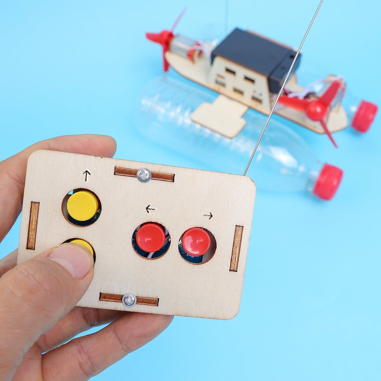 Hand holding a small wooden control panel with buttons, with a toy boat in the background on a blue surface.