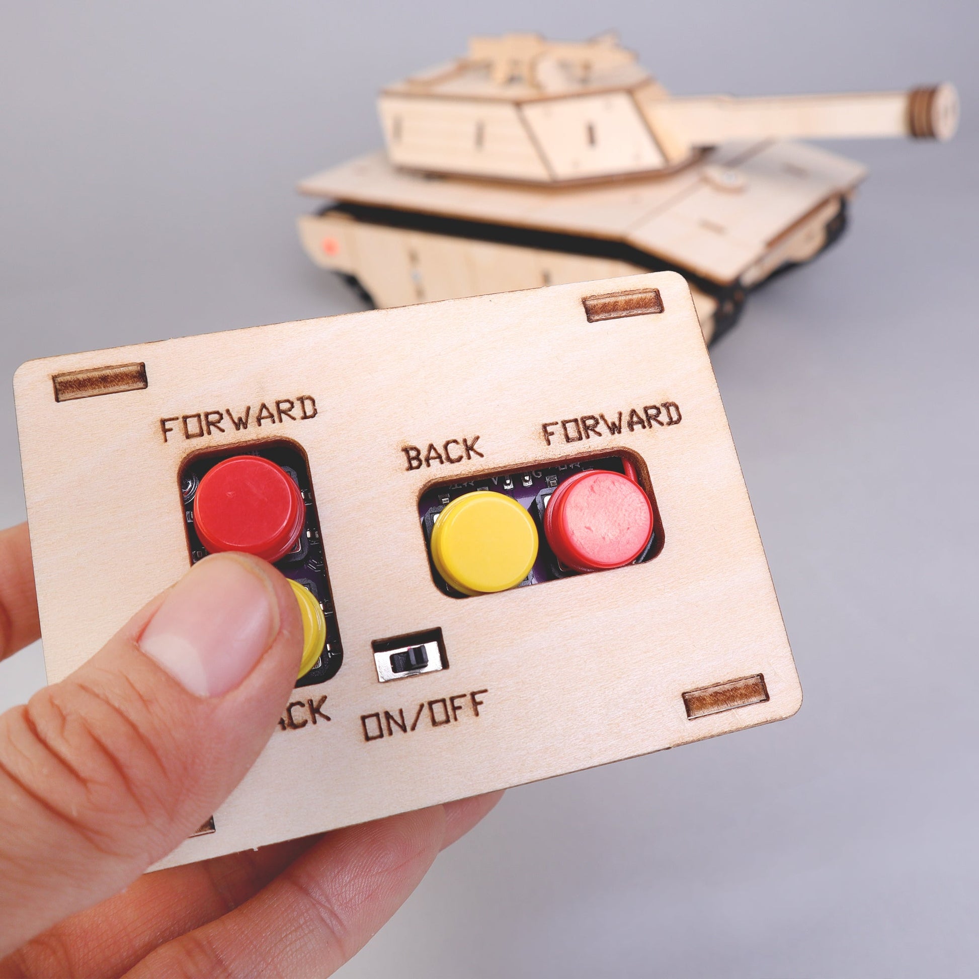 Wooden toy remote control with buttons held by a hand, with a toy tank in the background.