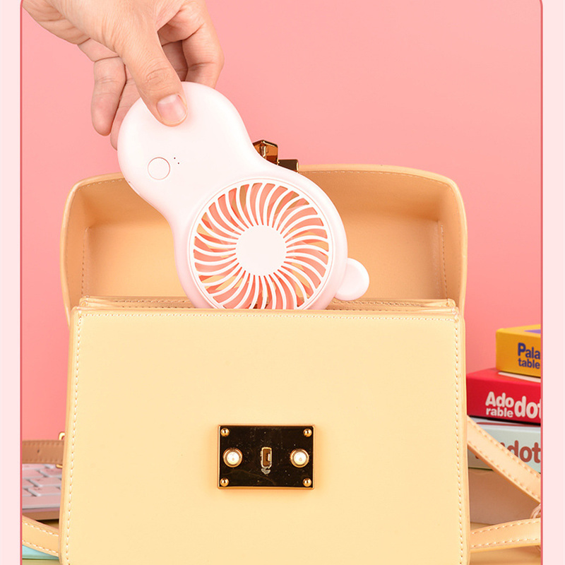 Hand placing a small white fan into a yellow handbag against a pink background