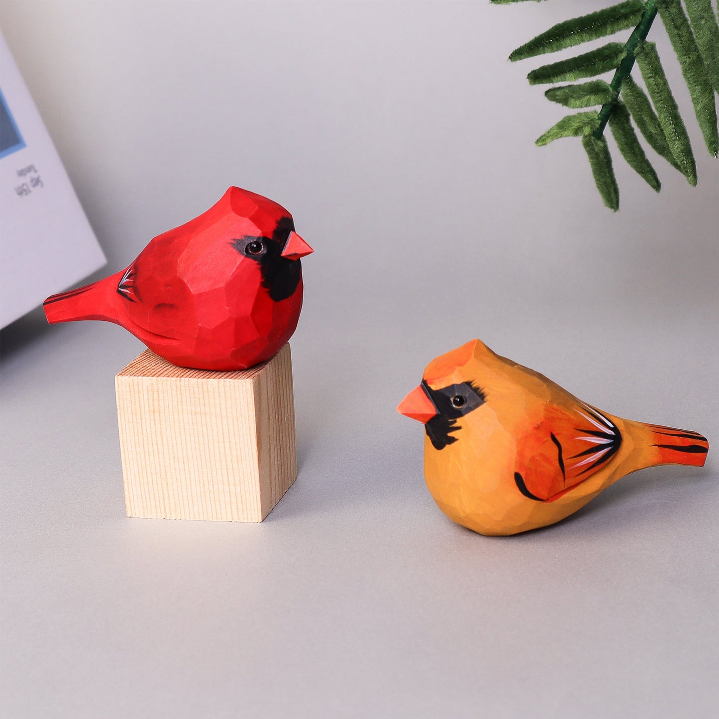 Hand Carved Cardinal Bird, one red and one orange, on a light gray surface with a plant in the corner.
