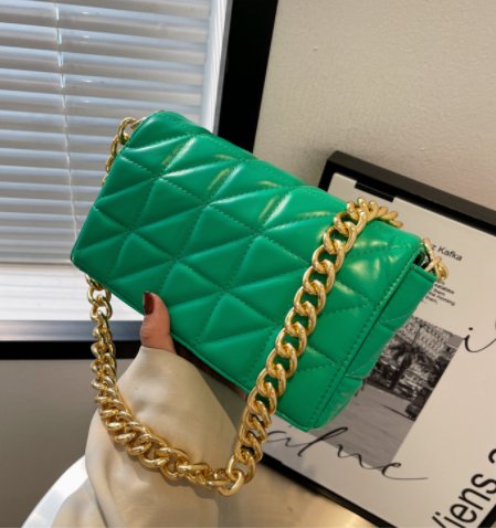 Green quilted handbag with gold chain held by a hand, with a blurred indoor background.