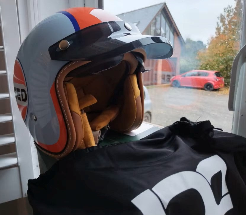 Motorcycle helmet with orange and gray design on a black jacket, with a car visible through a window in the background.