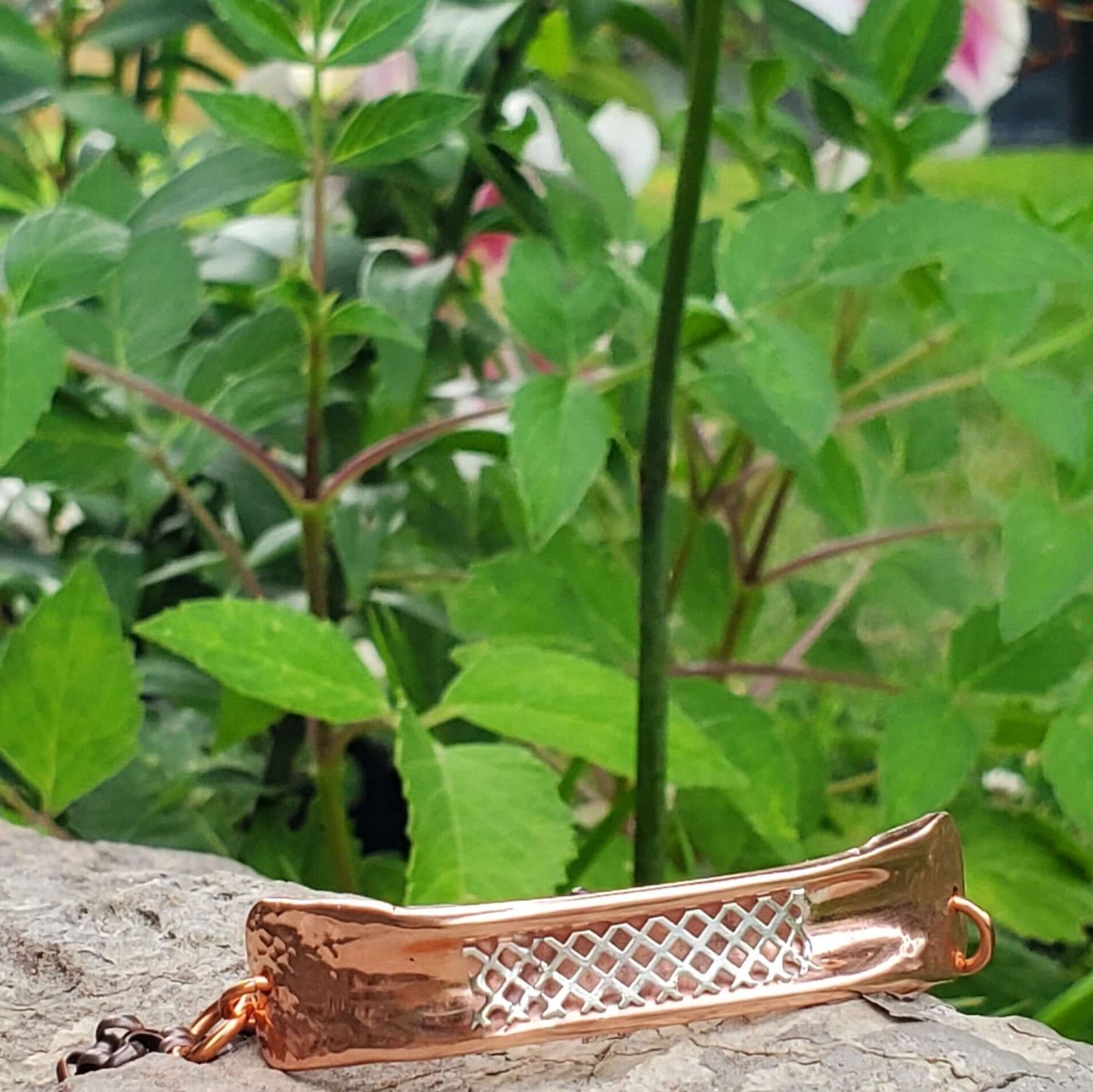 Copper-colored bracelet with intricate design on a rock with green foliage in the background