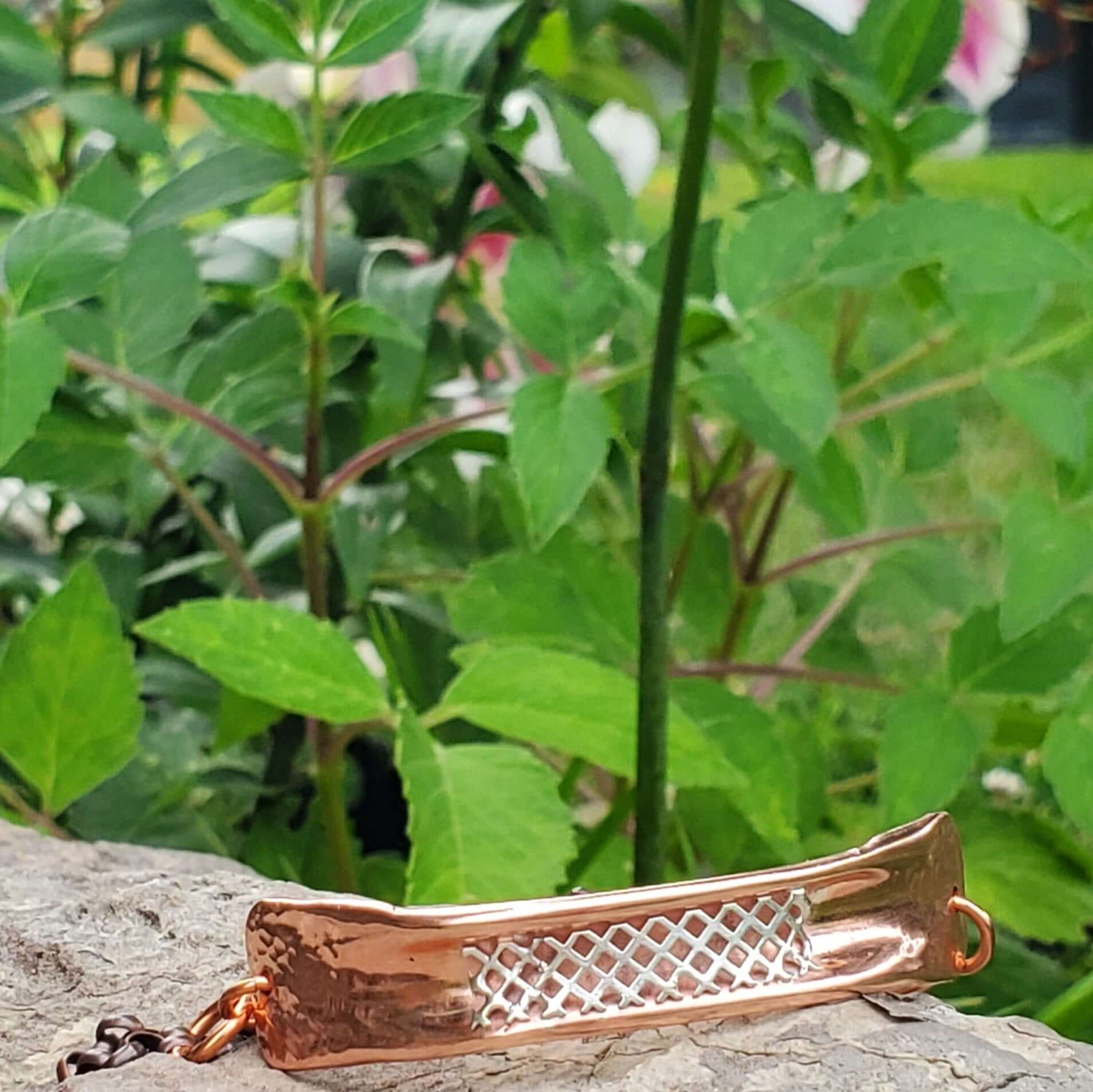 Copper-colored bracelet with intricate design on a rock with green foliage in the background
