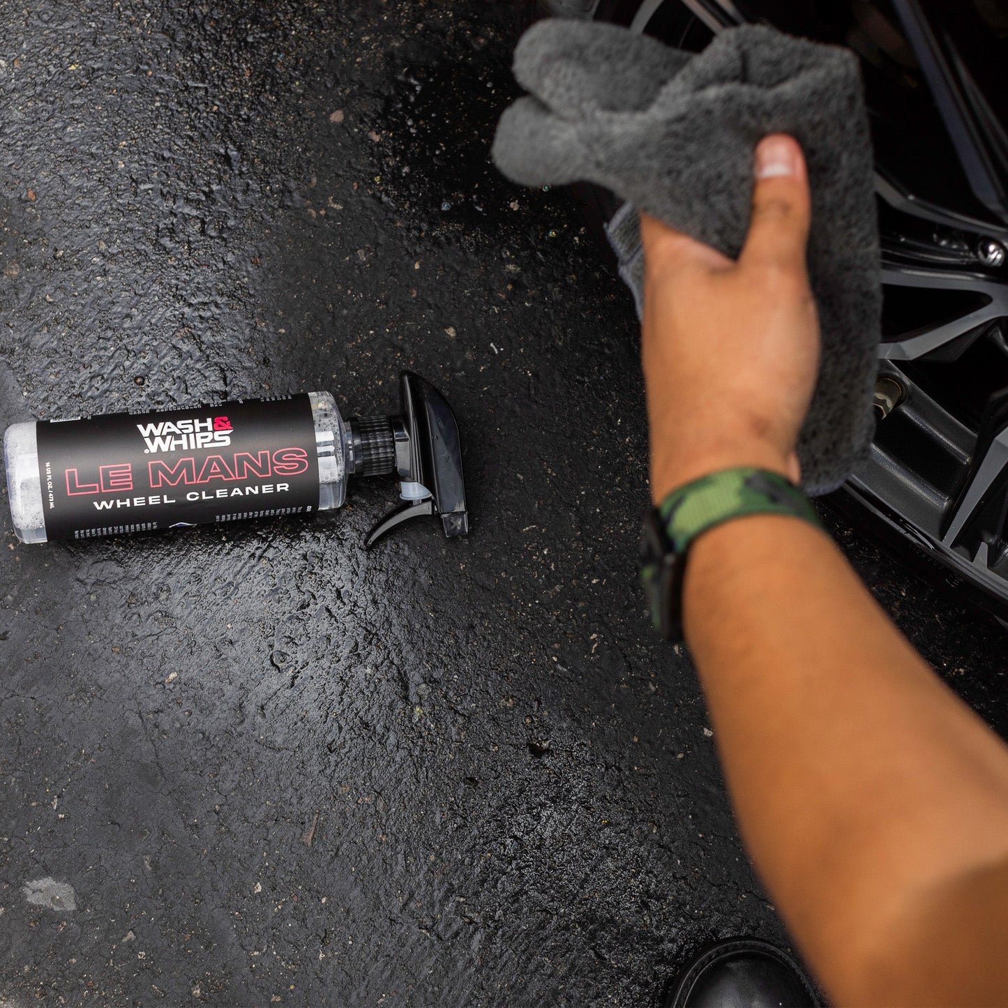Hand holding a gray sponge next to a tube of Wheel Cleaner on a black surface