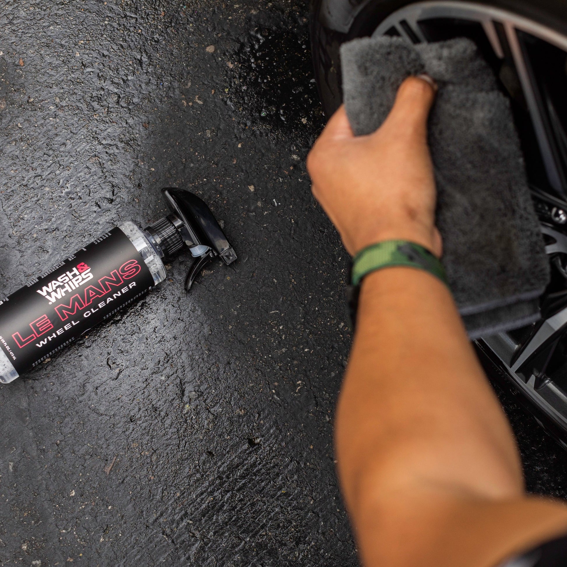 Person cleaning car wheel with a cloth and Le Mans wheel cleaner on a black surface