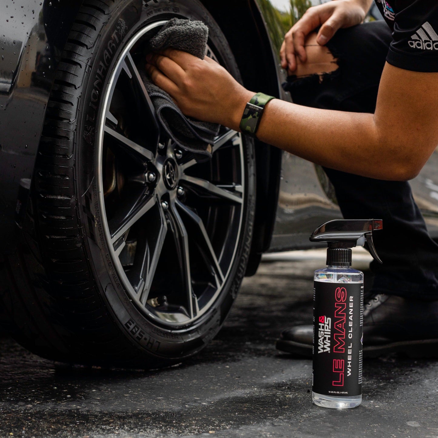 Person cleaning a car wheel with a spray bottle labeled 'Le Mans' on a dark surface.