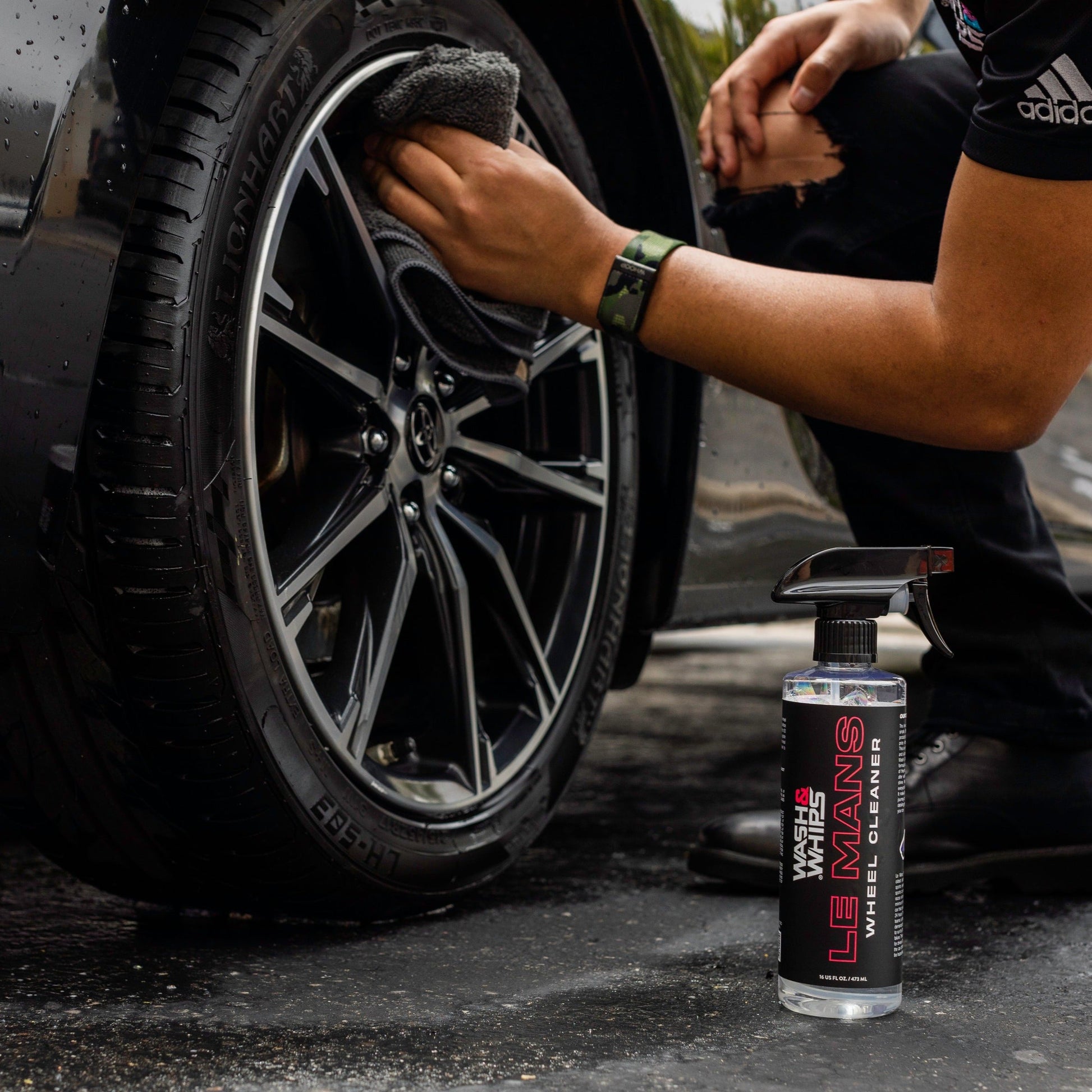 Person cleaning a car wheel with a spray bottle labeled 'Le Mans' on a dark surface.
