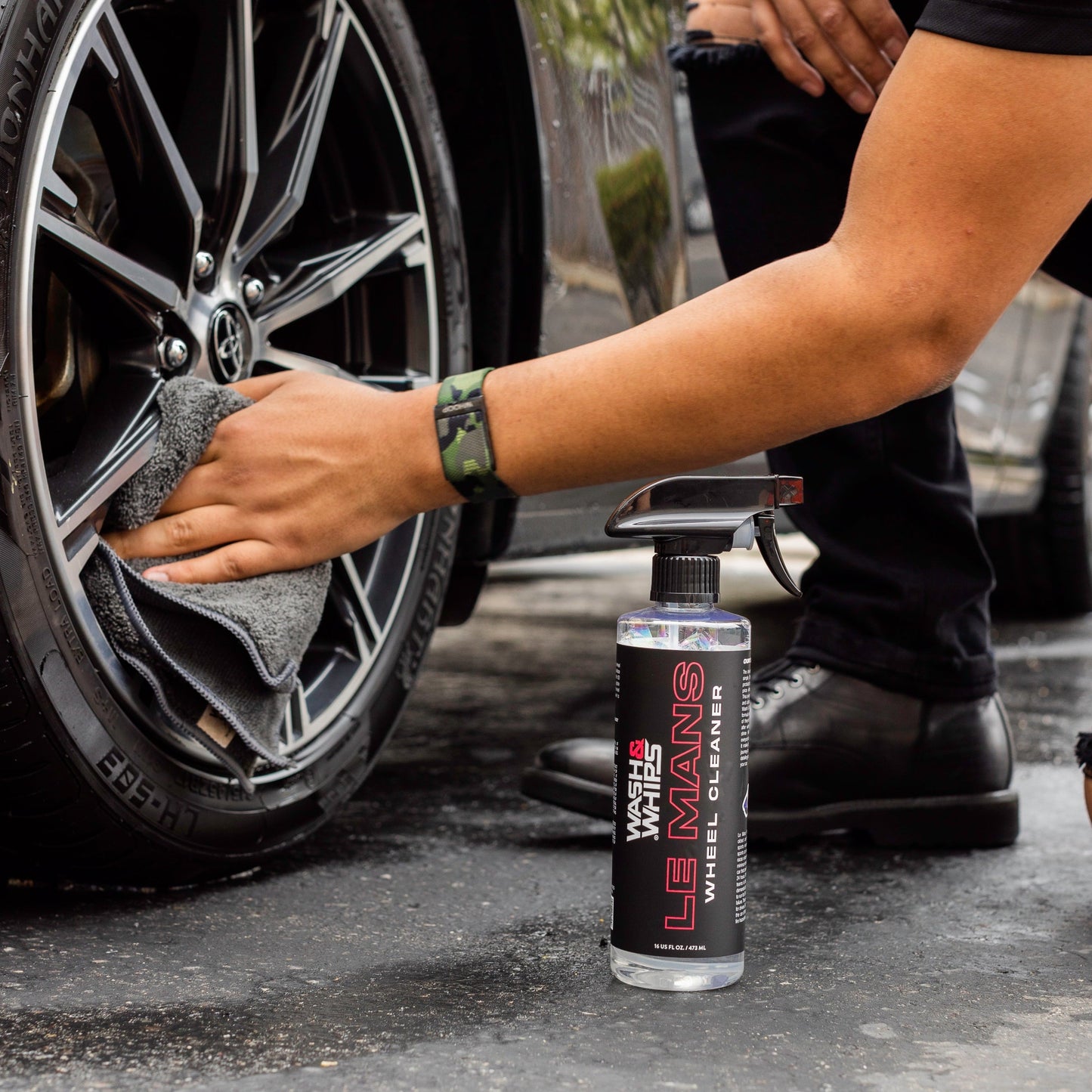 Person cleaning a car wheel with a spray bottle labeled 'Le Mans Wheel Cleaner' on a driveway.