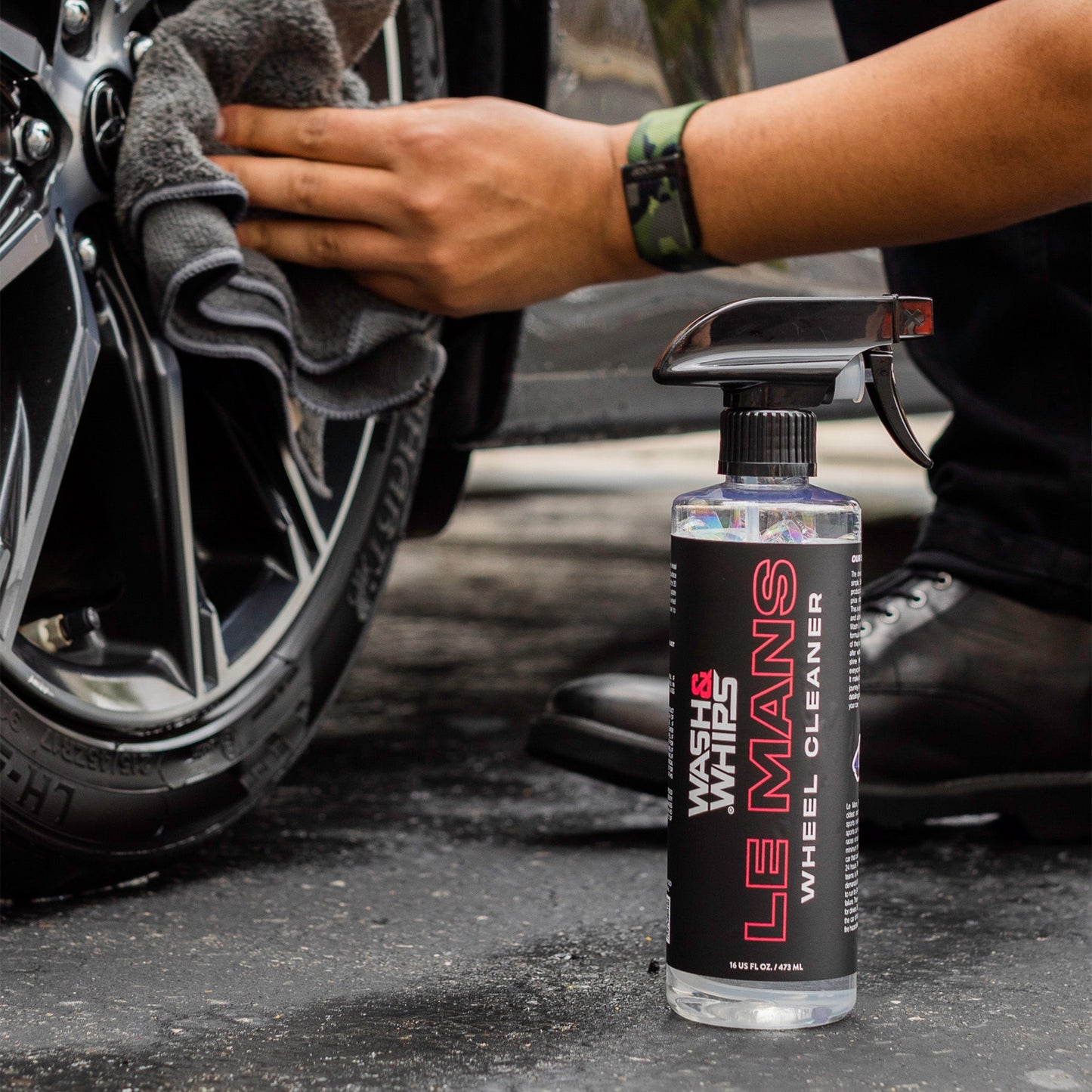 Person cleaning a motorcycle wheel with a towel, Le Mans Wheel Oil Cleaner spray bottle in foreground.