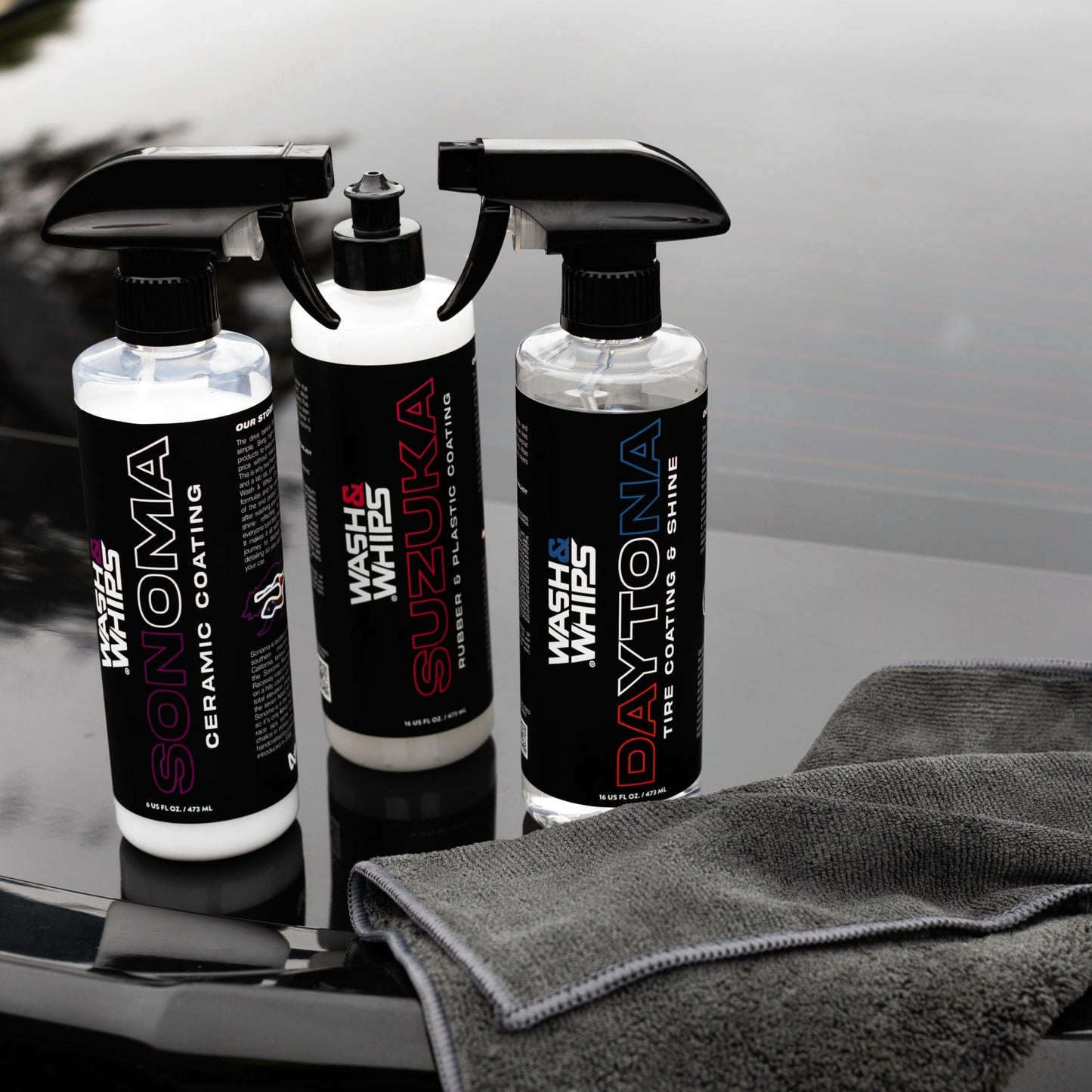 Three car care products with spray nozzles on a reflective surface, one of which is a gray towel.