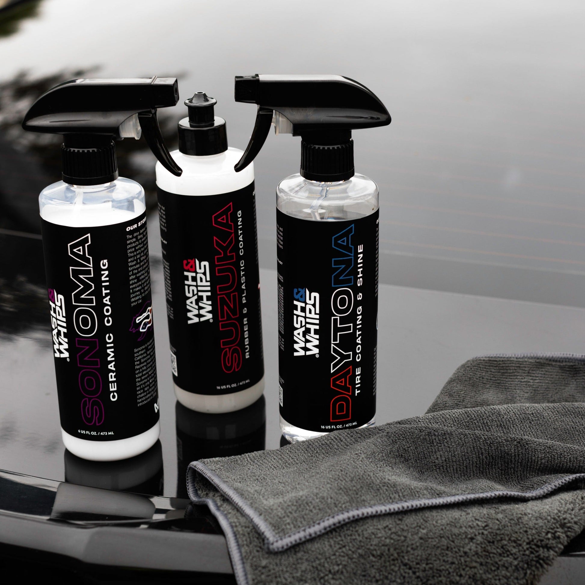 Three car care products with spray nozzles on a reflective surface, one of which is a gray towel.