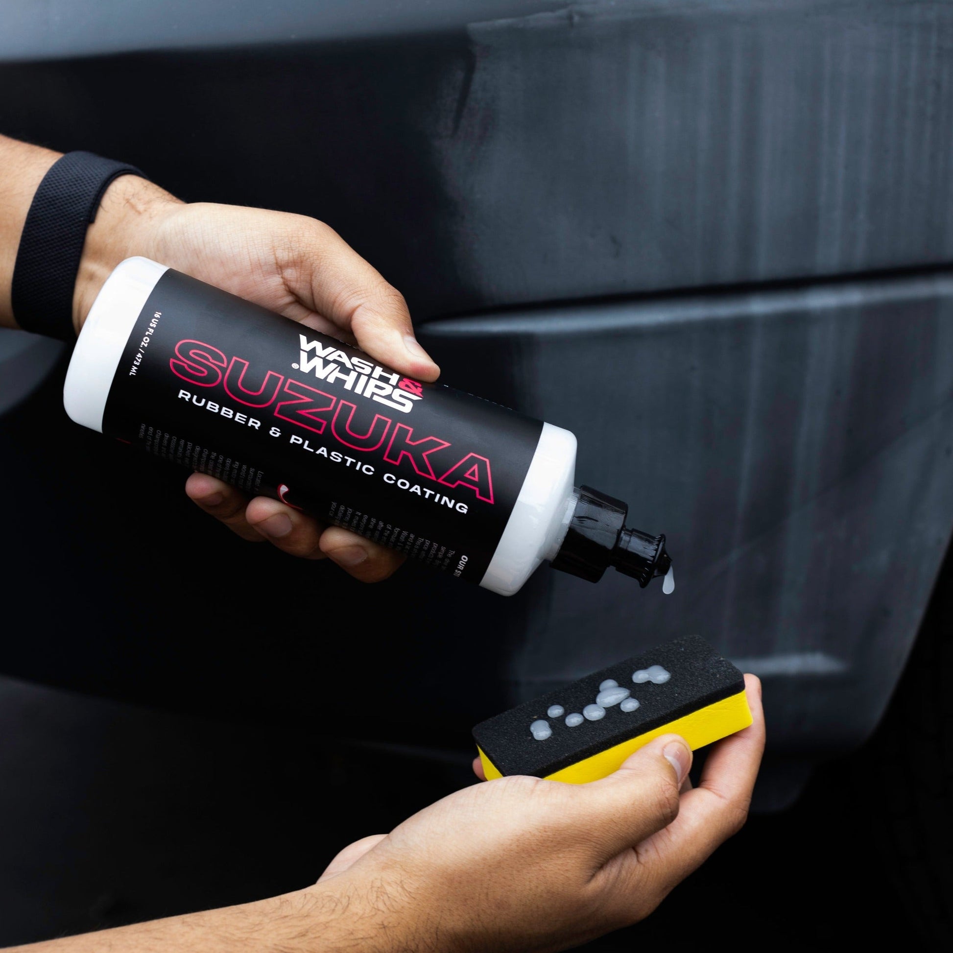 Person holding a bottle of 'Wash and Whips Suzuka' car care product with a sponge against a black car.