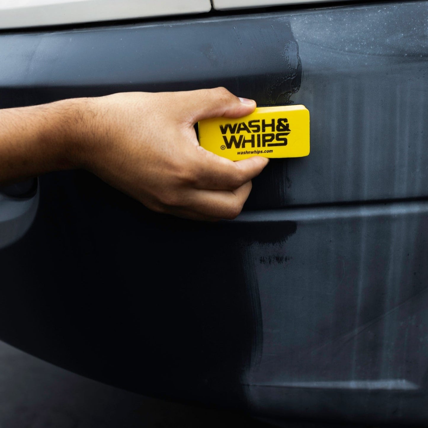 Person using a yellow 'Wash & Whips' product on a car door