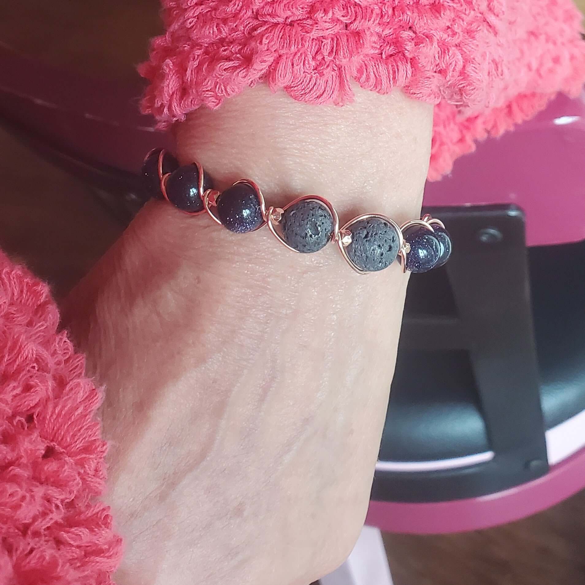 Close-up of a wrist wearing a bracelet with dark beads and rose gold accents, against a pink textured background.