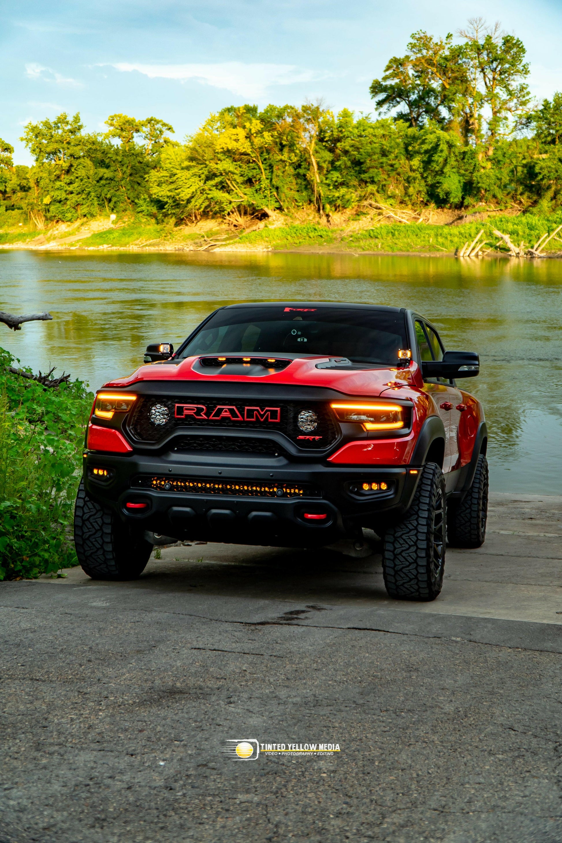 Red Ram truck with fog lights and light bar parked by a river with greenery in the background