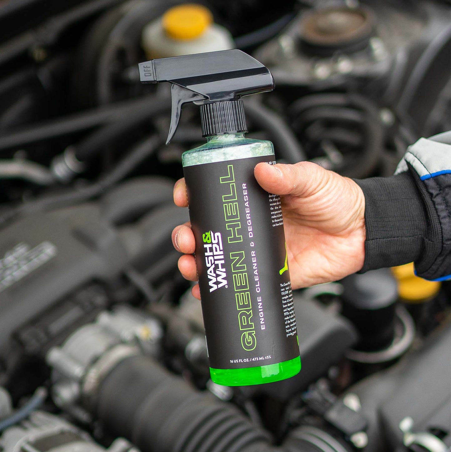 Hand holding a bottle of 'Green Hell' engine cleaner in front of an open car engine.