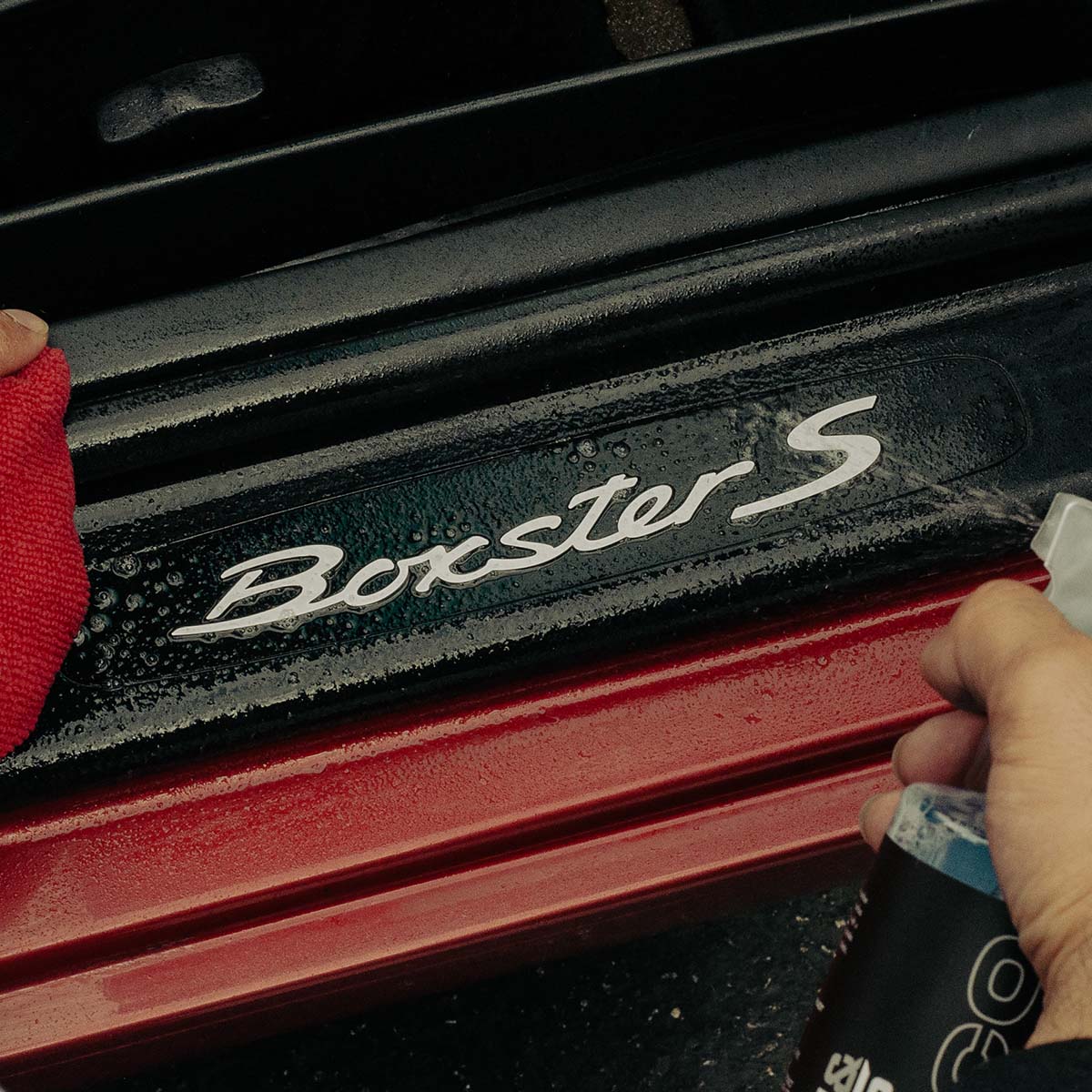 Person cleaning a Porsche Boxster S car with a cloth and spray bottle.